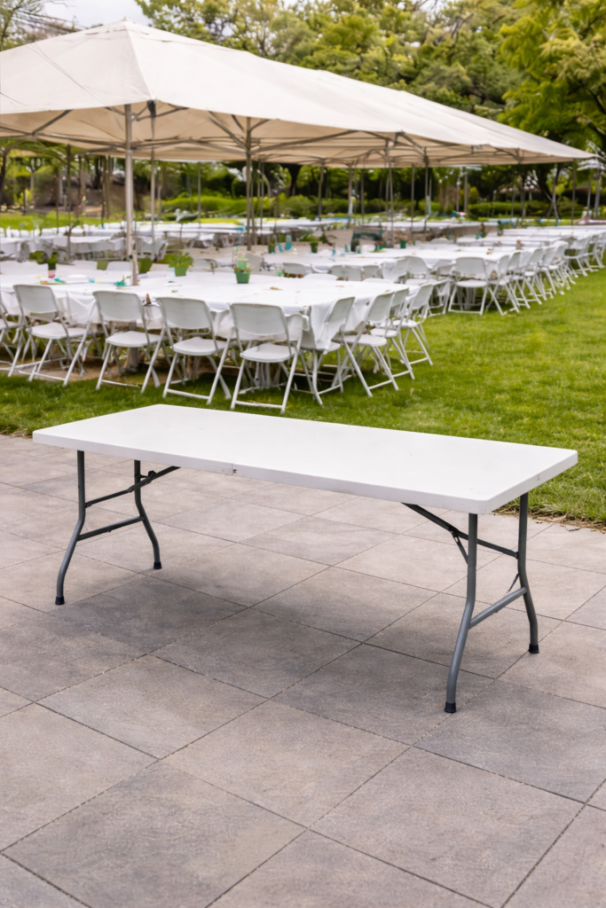 : Mesa plegable de catering para eventos culturales en exterior, con estructura metálica y tablero blanco bajo carpa.